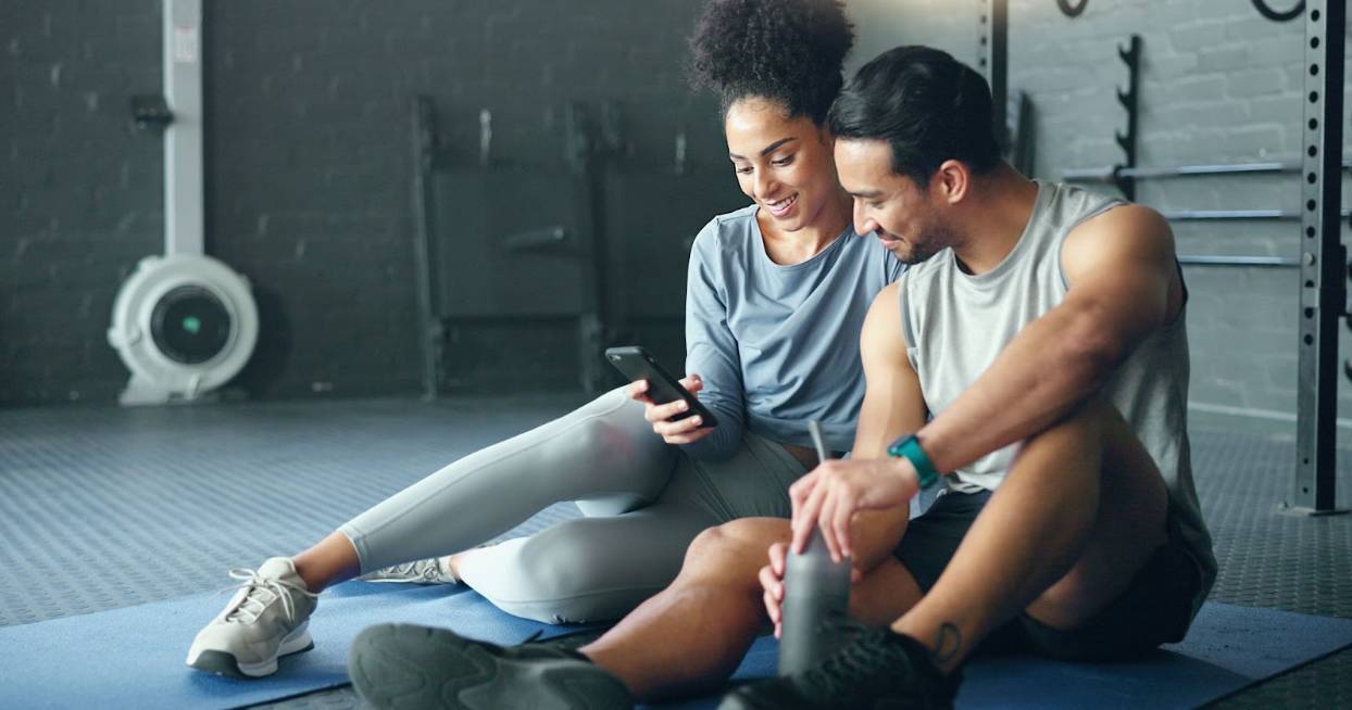 Two people sitting on gym mats after a workout, smiling and looking at a smartphone together in a fitness studio.