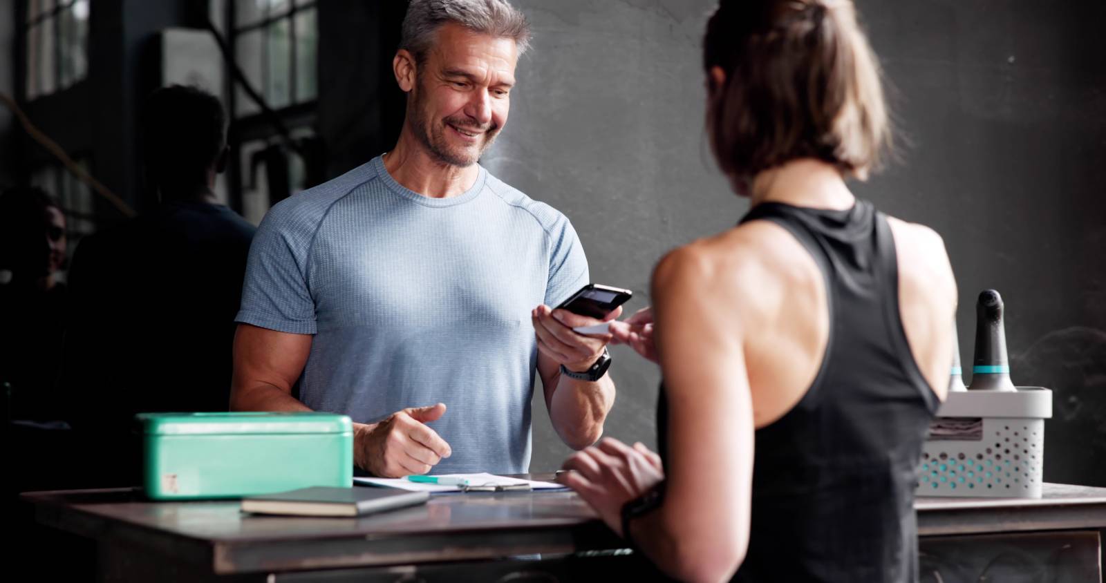 Gym staff member assisting a member at the front desk using a mobile device.